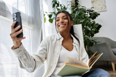 Mutlu genç bir kadın evde çalışıyor, akıllı telefonuyla meşgul oluyor ve elinde bir kitap tutuyor..