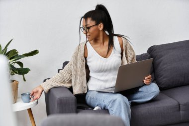 A young woman sitting on a couch, engaged in studying with a laptop and sipping coffee. clipart