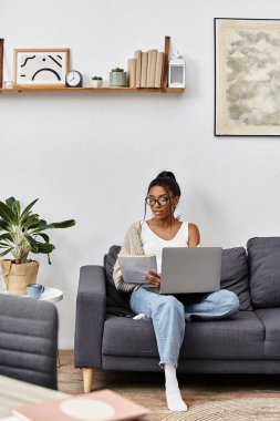 Genç bir kadın konforlu, modern bir oturma odasında laptopuyla rahatça ders çalışıyor..