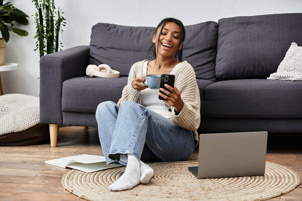 A young woman relaxes on the floor, studying with a laptop, coffee in hand, surrounded by comfort.