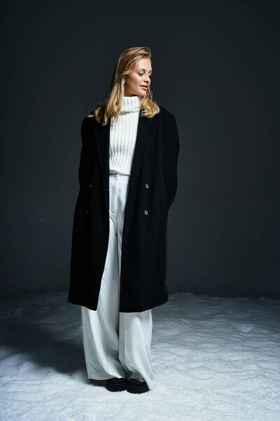 Молодая женщина в шикарном черном пальто и белом наряде уверенно стоит на тёмном фоне.