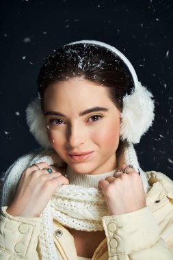 A woman smiles warmly while wearing ear muffs and a cozy scarf against falling snowflakes. clipart