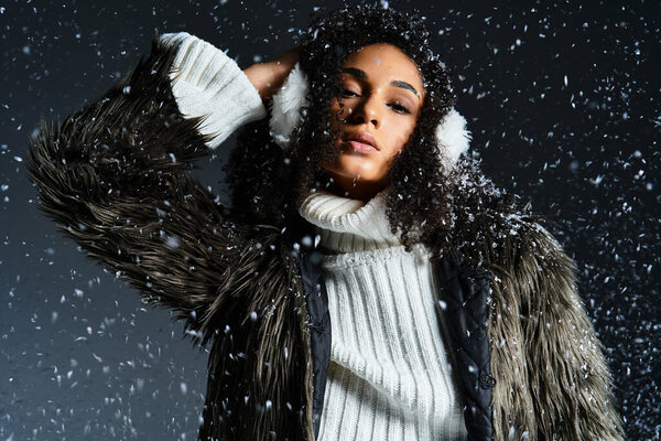 A stylish brunette showcases winter apparel with flair, wearing earmuffs and a warm sweater amidst falling snow.