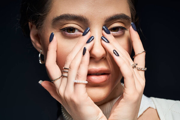 A graceful woman gently touches her face, showcasing stunning nail art and accessories.