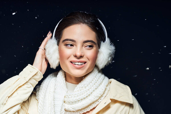 The woman smiles warmly while snow gently falls around her in a cozy winter setting.