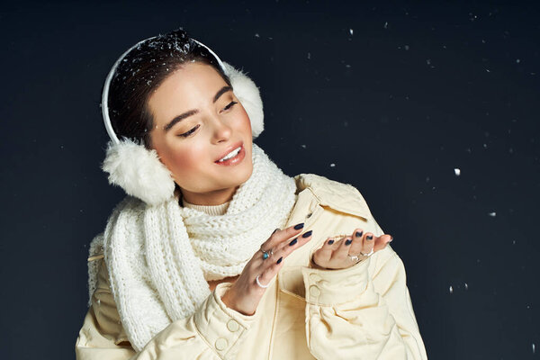 Улыбающаяся женщина играет с падающими снежинками, надевая теплые наушники и шарф.