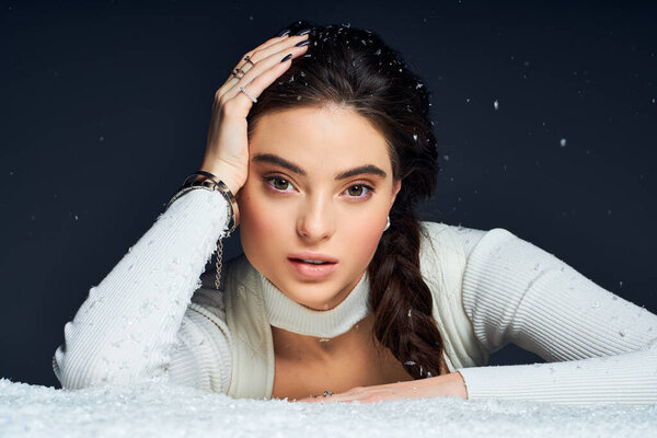 A stunning woman with a braided hairstyle rests her hand on her head, surrounded by snowflakes.