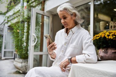 Güzel, olgun bir kadın açık havada dinleniyor, telefonuna dalmış, canlı çiçeklerin arasında..