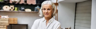 Neşeli, olgun bir kadın sanat atölyesinde gururla duruyor. Etrafı benzersiz el sanatlarıyla çevrili..