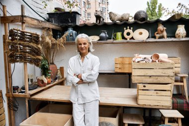 Kendine güvenen olgun bir kadın, etrafı çömlek ve dekorla çevrili sanat atölyesinde duruyor..