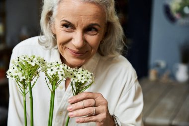 A joyful mature woman enjoys the fragrance of beautiful flowers in her hands, radiating happiness. clipart