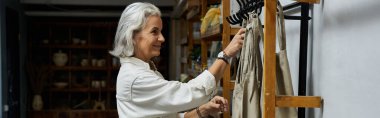 Güzel, olgun bir kadın şık bir butik ortamında kıyafet düzenlerken gülümsüyor..