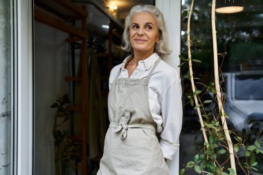Neşeli olgun bir kadın pencereye yaslanır, temiz havanın ve doğal ışığın tadını çıkarır..
