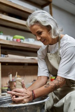 A beautiful mature woman skillfully shapes clay while smiling in her pottery studio. clipart