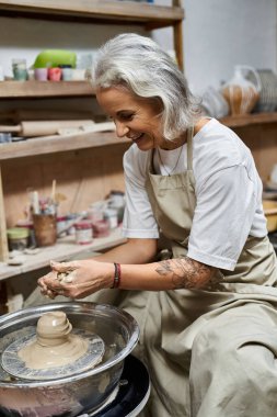 A skilled mature woman joyfully shapes clay on a pottery wheel in her art studio. clipart