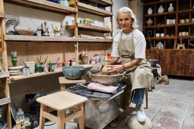 A talented mature woman expertly molds clay in her inviting pottery studio, surrounded by tools.