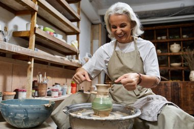Yetenekli bir zanaatkar, aletlerle çevrili rahat çömlek atölyesinde gülümserken kili şekillendirir..