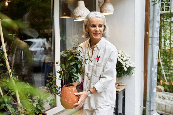 A joyful woman with silver hair carries a potted plant, smiling in a garden shops outdoor space.