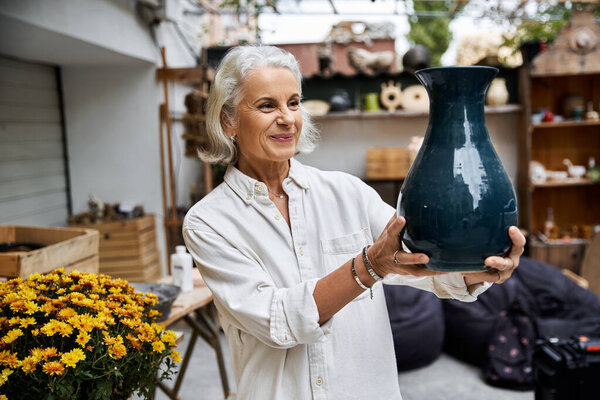 A joyful mature woman presents a striking pottery vase surrounded by lush greenery.