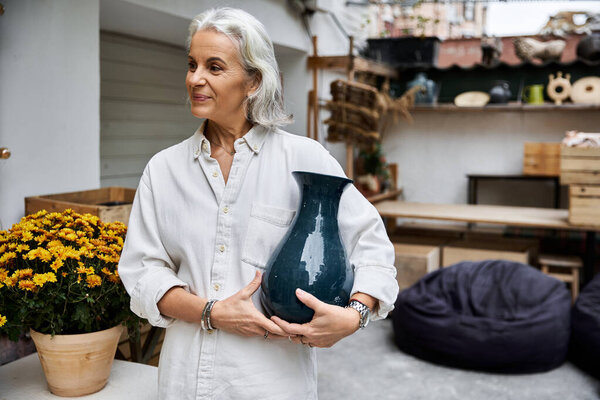 A graceful mature woman holds a stunning vase while surrounded by artistic decor and flowers.