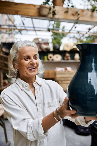 A mature woman smiles brightly while holding a stunning piece of pottery in her studio.