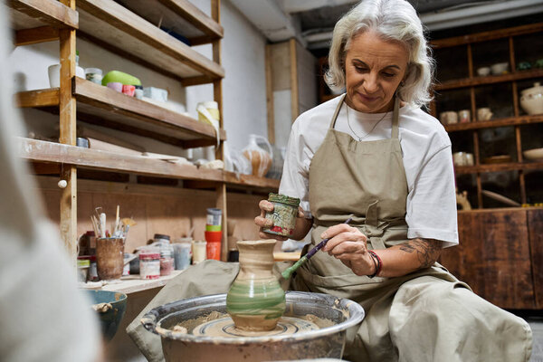 A talented mature woman meticulously decorates a handmade pottery piece in her bright studio.