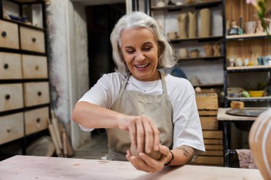 A beautiful mature woman joyfully shapes clay while immersed in her creative pottery studio. clipart