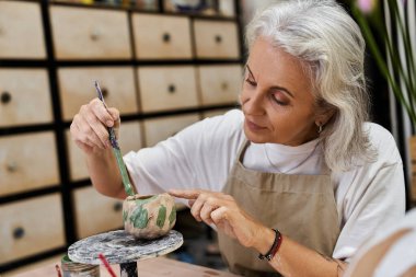 A skilled mature woman focuses on adding details to her pottery project in a vibrant studio. clipart