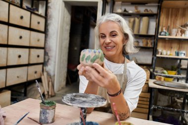 A skilled artisan proudly displays her handmade pottery while surrounded by creative tools. clipart