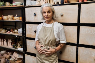 A beautiful mature woman stands proudly in her pottery workshop, crafted pieces all around her. clipart