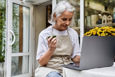 The woman sips tea from a cup while focused on her laptop in a tranquil outdoor space. clipart