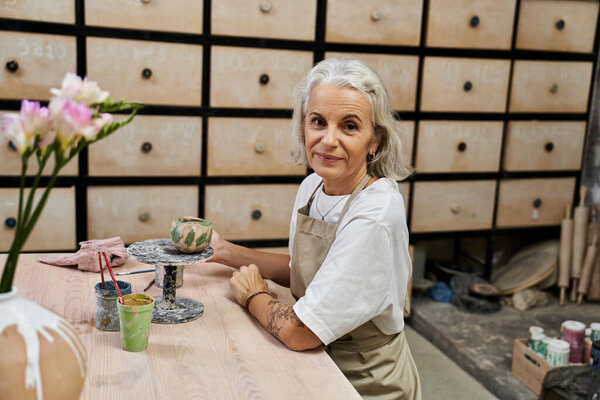 Зрелая женщина занимается керамикой, в окружении художественных материалов и хранения.
