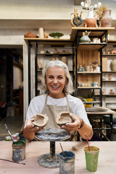 A smiling mature woman showcases her handcrafted pottery in a vibrant art studio.