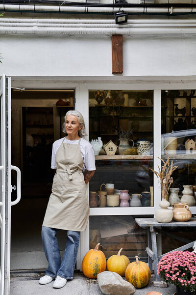A graceful mature woman leans against the entrance of her pottery shop surrounded by seasonal decor.