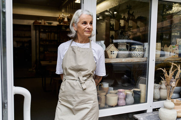 The mature woman warmly welcomes customers to her inviting pottery shop filled with unique pieces.