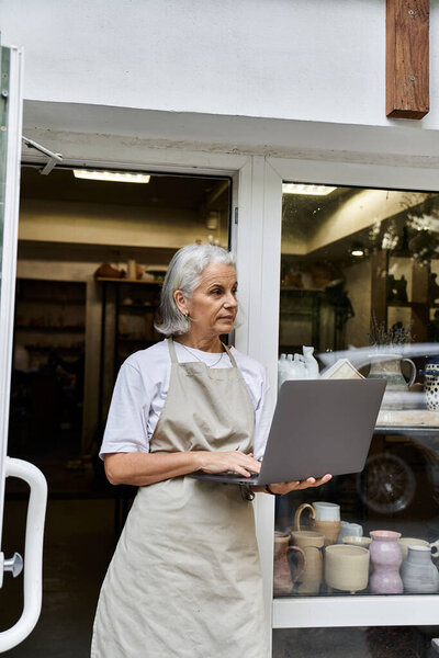 A beautiful mature woman types intently on her laptop while standing at her shop entrance.