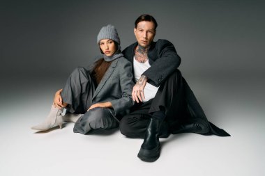 A young couple dressed in fashionable attire sits together on the floor in a contemporary studio.