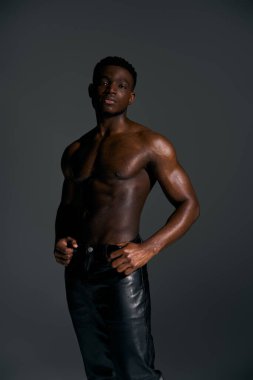 Genç, yakışıklı, Afro-Amerikan bir adam kendine güvenen üstsüz bir şekilde duruyor, koyu renk minimalist bir arka plana karşı tonlu fiziğini vurguluyor. Davranışları kendine güven ve karizma yayıyor..