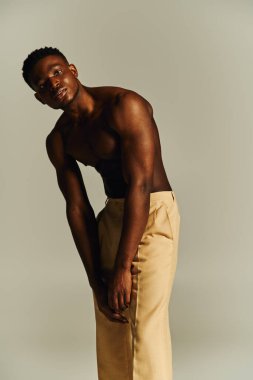 A young handsome African American man stands confidently, posing topless while wearing tailored beige pants. His posture exudes elegance and self assurance, set against a minimalist backdrop. clipart