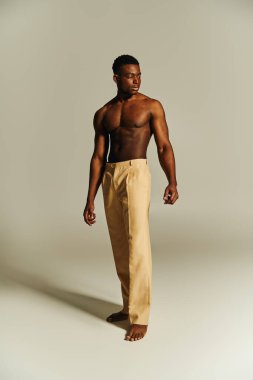 A young handsome African American man stands confidently, showcasing his toned physique. Clothed in beige pants and barefoot, his pose reflects strength and style amidst a plain background. clipart