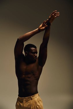Genç, yakışıklı bir Afro-Amerikan erkek, fiziğini göstererek poz veriyor. Dik duruyor, kollarını kaldırıyor, iç mekanda güç ve sanatsal bir yapı oluşturuyor..
