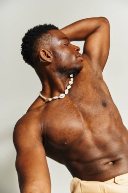 A young African American man stands confidently, showcasing a sculpted physique while wearing a unique necklace. The studio background enhances the focus on his powerful pose and vibrant energy. clipart