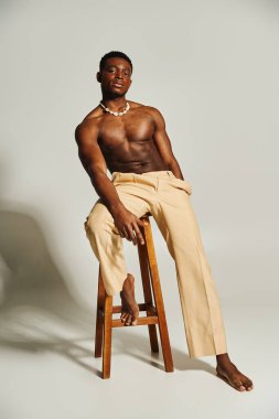 Kendine güvenen Afro-Amerikan bir adam tahta bir taburede üstsüz poz veriyor..