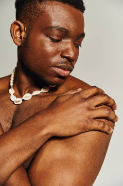 Genç ve çekici bir Afrikalı Amerikalı üstsüz poz veriyor, fiziğinin tonunu vurguluyor. Nazikçe bakıyor, eşsiz kolyesini yumuşak, tarafsız bir arka plana doğru gösteriyor..