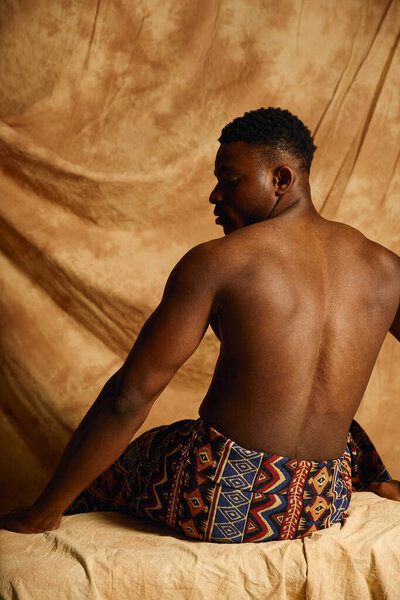 A young handsome african american man poses topless, exuding confidence and charm. He is seated on a textured surface, dressed in a patterned wrap, with a neutral backdrop enhancing his features.