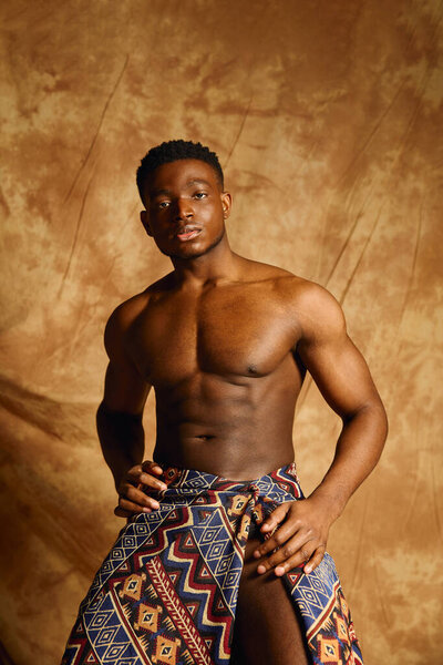 A youthful and attractive African American man presents a powerful stance without a shirt. He wears a colorful wrap and stands against a textured brown backdrop, exuding confidence and charisma.