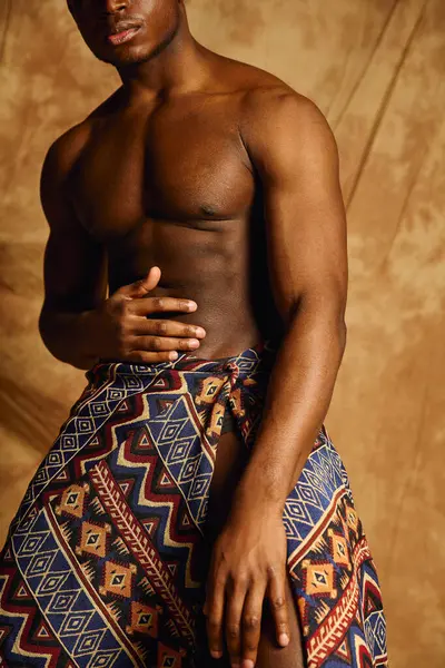 Genç ve yakışıklı Afrikalı Amerikalı erkek, sanatsal bir ortamda üstsüz poz verirken fiziğini sergiliyor. Sıcak arka plan, sargısının canlı desenlerini güçlendiriyor ve kendine güveni yayıyor..