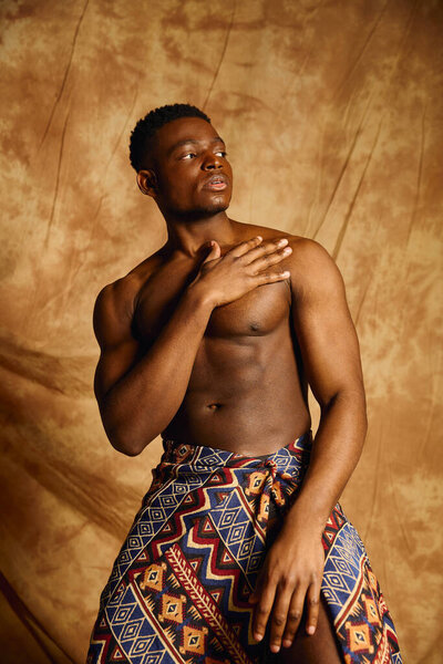 A young, handsome African American man showcases his physique, posing topless with elegance. Draped in vibrant tribal fabric, his expression exudes confidence and charisma in this artistic setting.