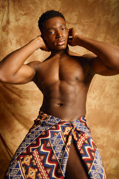 A young handsome African American man poses confidently, showcasing his physique. Wearing a patterned wrap, he embodies strength and culture against a warm, artistic backdrop.