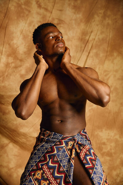 A young handsome African American man, showcasing his physique, stands confidently with his hands on his neck. He wears a traditional patterned wrap, exuding charm and charisma in an artistic setting.